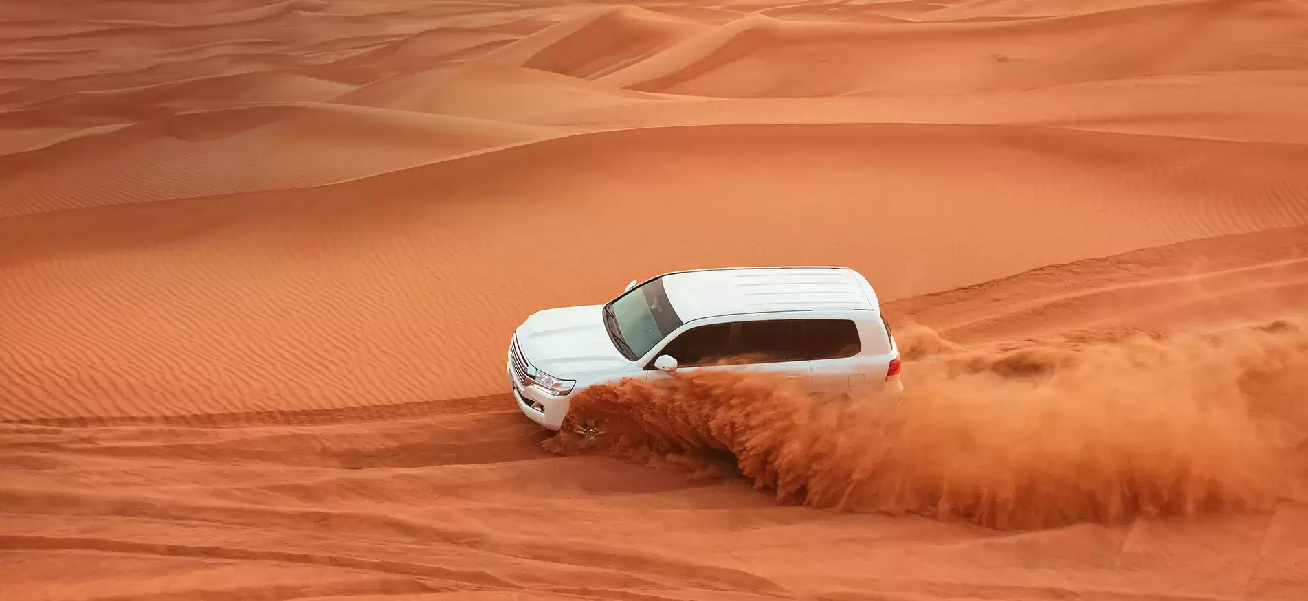 White SUV driving fast across red desert sand dunes, kicking up a large cloud of dust